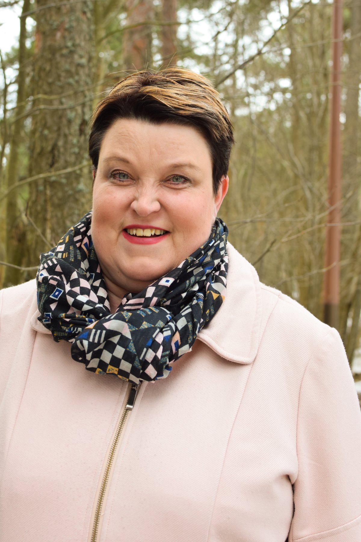 Nainen kurkistaa hymyillen kahden ison koivun välistä keltaisessa takissaan.